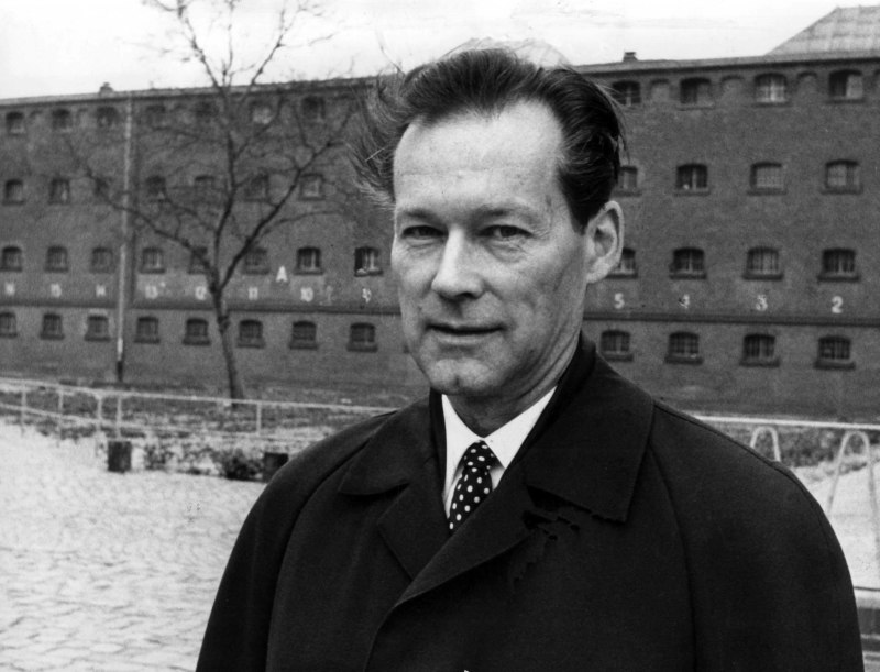 A black and white photo of a dark-haired white man in a dark jacket and polka-dot necktie. He is standing outside the prison smiling at the camera.