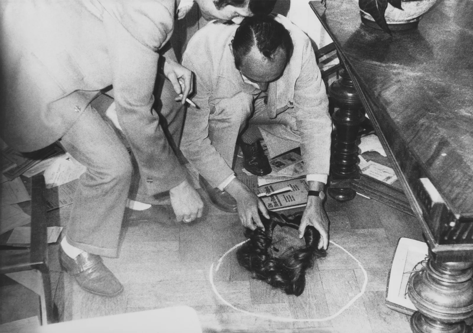A black and white photo shows two men in suits bending down toward a discarded wig lying in a chalk circle on the floor.