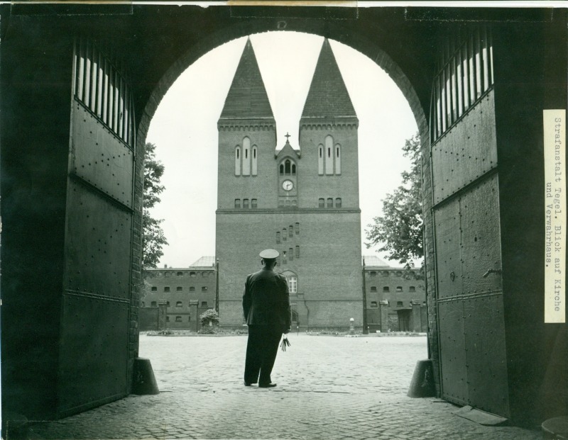 A black and white photo of a cobblestone courtyard seen through the open gate in the outer wall. At the far end of the courtyard is a multi-story chapel with two spires. A uniformed guard stands facing away from the viewer.