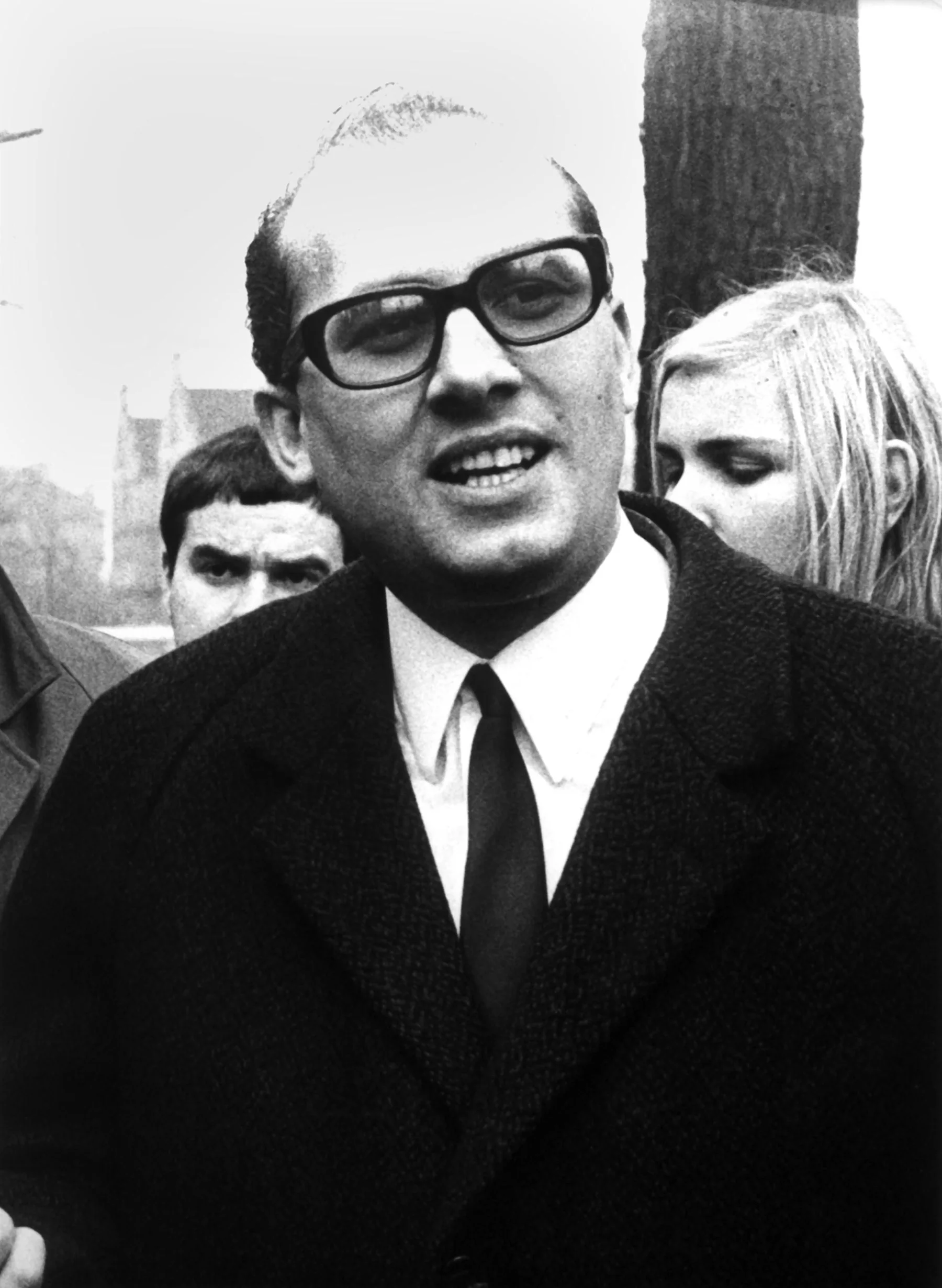 A black and white photo of a balding white man wearing thick black glasses and a suit and tie. He is standing in front of several other people and looks as though he is in mid-sentence.
