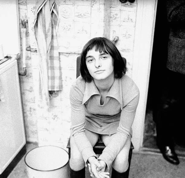 A black and white photo of Goergens, a young white woman with chin length straight dark hair and uneven bangs wearing a mini dress and knee high boots. She is sitting on a chair in a kitchen looking up at the viewer with an amused expression.