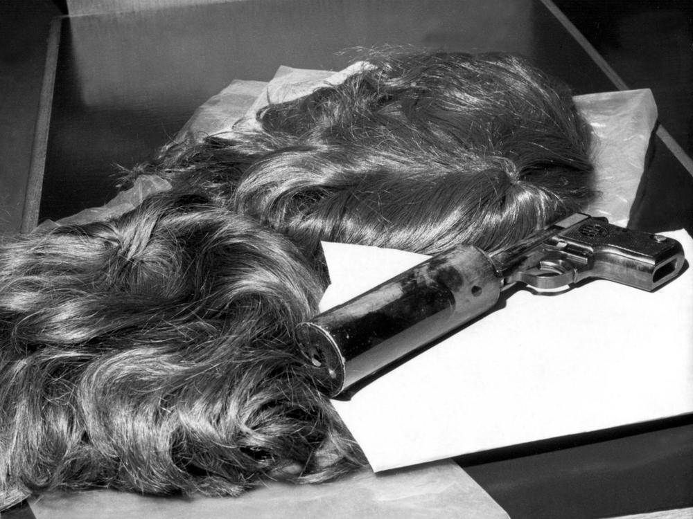 A black and white photo shows two poor quality wigs and a handgun with a silencer, each resting on a folded sheet of paper.