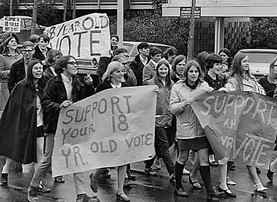 A black and white photo of a group of young men and women marching down a street carrying handmade signs supporting lowering the voting age to 18.