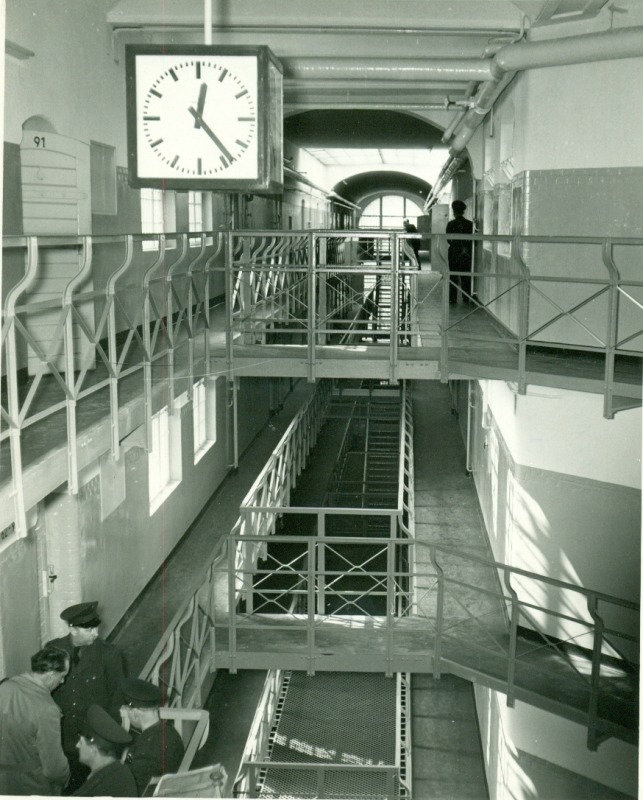 A black and white photo of a the staircase and hallway of a multi-story prison block. The upper storeys do not cover the whole floor but have large gaps in the middle with narrow pathways hugging the walls. The stairs and pathways are surrounded by metal pipe railings. At the bottom left of teh photo, three prison guards surround a prisoner.