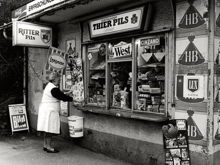 A black and white photo of the front window and counter of a small newsstand, covered in advertising posters for beer and newspapers.  An elderly woman in a white smock adjusts a poster to the left of the window.  Inside the building a jumble of products for sale is visible.