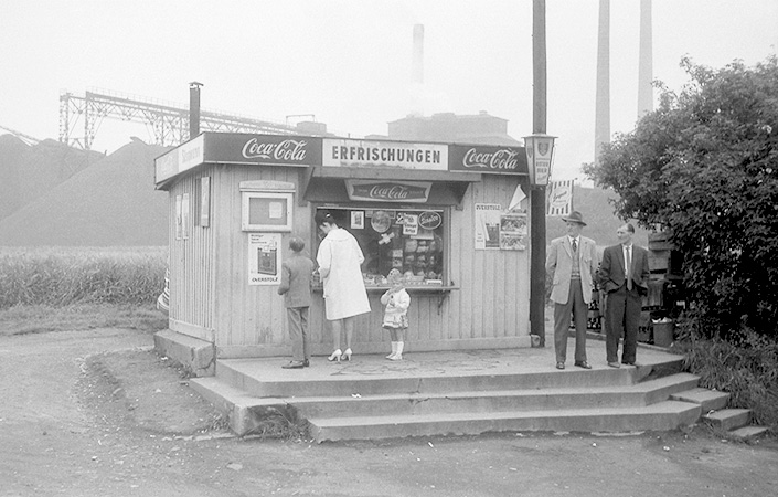 A black and white photo of a very small wooden newsstand about the size of a garden shed, in the middle of an empty lot. A woman with her two children stands at the window.  On the opposite side of the building, two men in business suits stand at the top of the steps leading away from the newsstand, looking toward the woman.