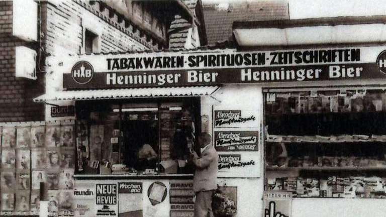 A black and white photo of a concrete newsstand next to a taller brick building.  A man in a business suit stands at the window speaking to the person inside.  Behind him, shelves are stocked with magazines and food items.