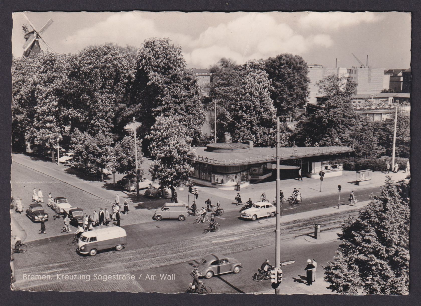 A black and white photo of a single-story modernist building with rounded corners at the intersection of two tree-lined streets.  Cars, cyclists, and pedestrians are passing by on the street and sidewalk. An old windmill is visible above the trees in the top left of the frame, while more modern multi-story buildings are visible to the right.