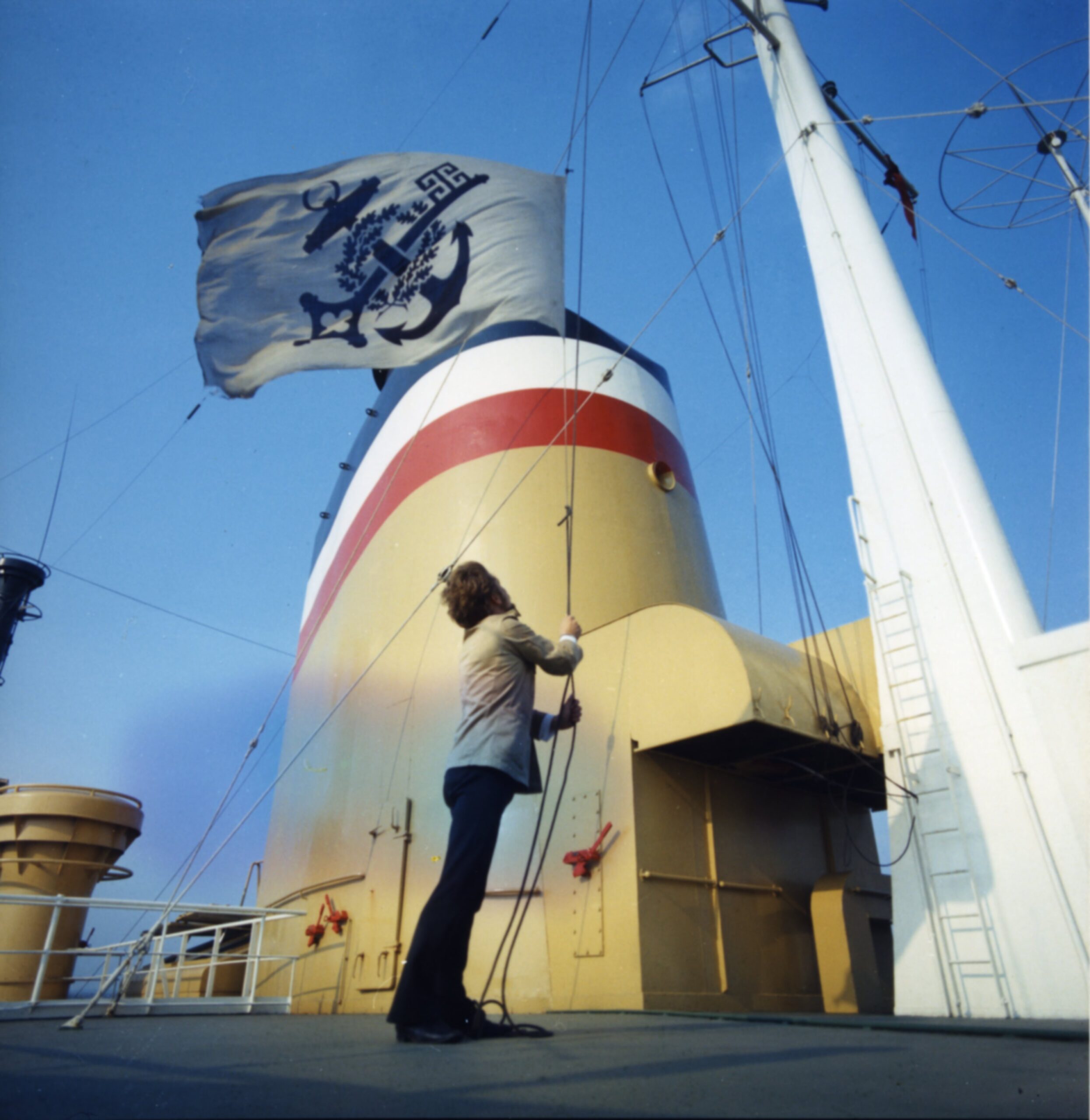 A color photograph of the upper deck of a ship against a bright blue sky.  A man in a white jacket and black pants stands in front of a yellow smokestack which is about twice his height.  Most of it is painted yellow.  At the top three stripes are painted in blue, white, and red.  The man is raising a white flag with a blue symbol on it which shows a ship's anchor crossed with an old-fashioned skeleton key, with a wreath woven around the crossing point. The flag's rope is connected to a white mast pole to his right.