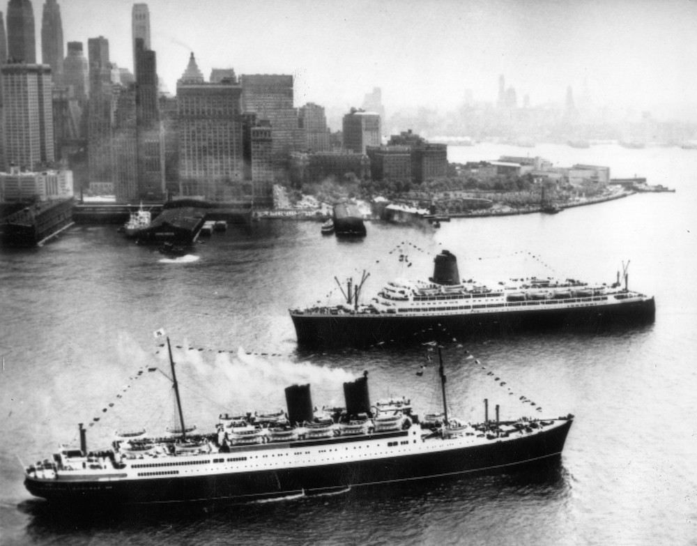 A black and white photo of two passenger ships, the MV Europa and the MV Bremen, on the river with New York skyscrapers on the shore behind them.  The ships are decorated with strings of small flags which attach at the bow and stretch upward across the tops of the two masts and then down to the stern. smoke rises from the smokestack of the nearer ship, but the calm water shows that they are not currently underway.