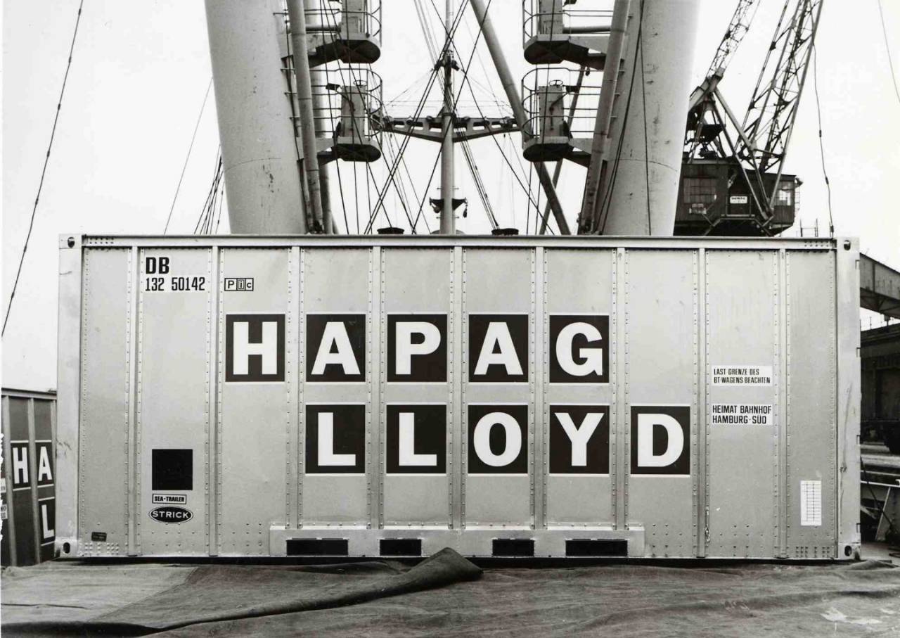 A black and white photo of a shipping container sitting on tarps on the deck of a ship.  Crane scaffolding and mast poles with myriads of cables stretched in multiple directions ascend out of the frame behind it.  The container is made of ten sheets of metal connected along the vertical edges with rivets.  Large black stickers with white letters have been placed on the middle six panels, forming the company name Hapag Lloyd. 