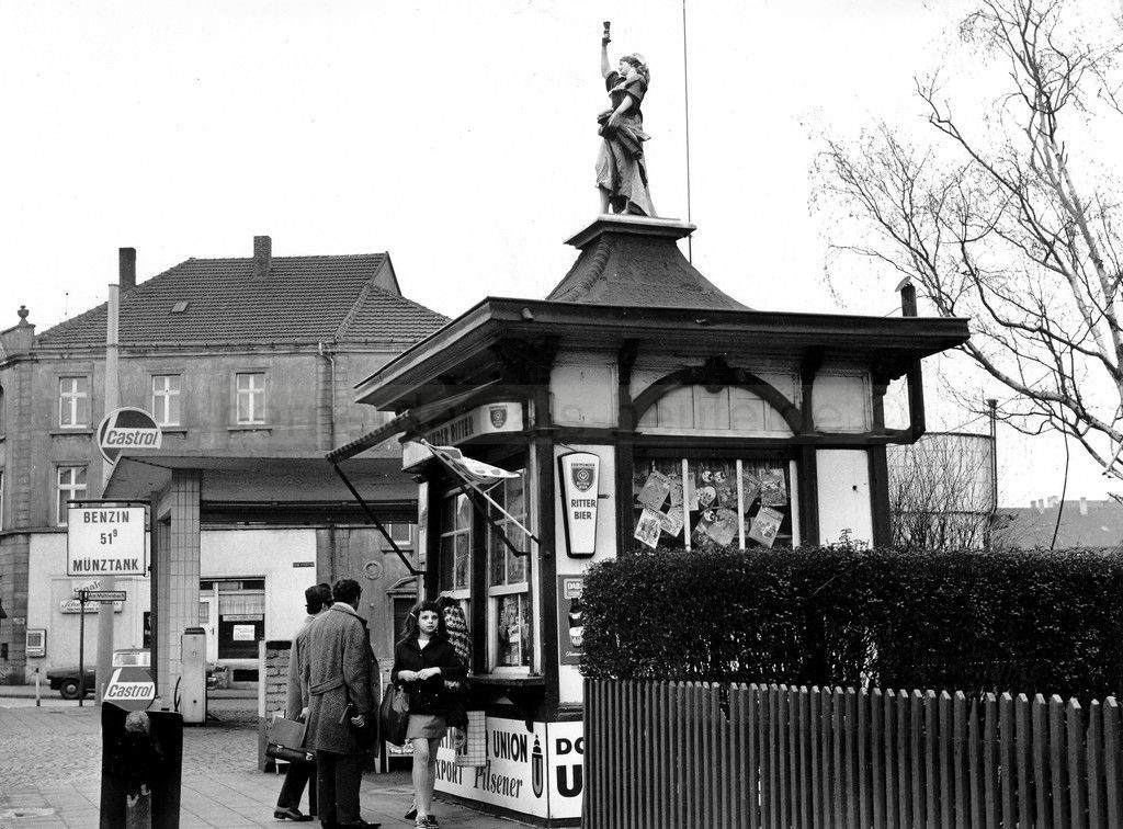 A black and white photo of a small newsstand decorated to look like a half-timber building, with a statue on the roof of a woman in long skirts holding up one arm as though raising a toast.  Several people wait in line at the window.