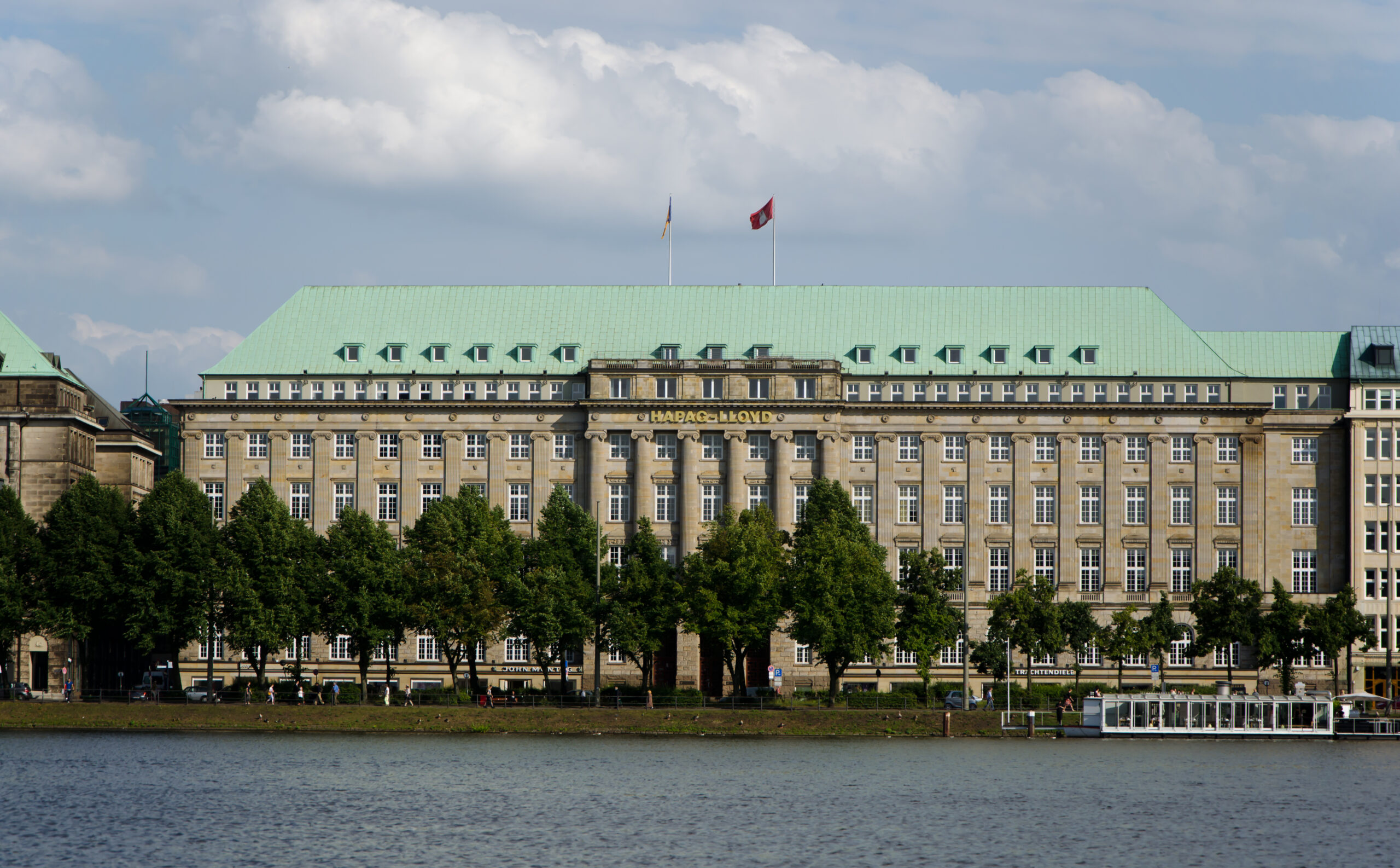 A color photograph of a multistory neoclassical building with long rows of four-paneled windows, a green roof and tan stone walls, sitting behind a neat row of trees along a riverbank.  Two flags fly from flagpoles at the top center of the building.
