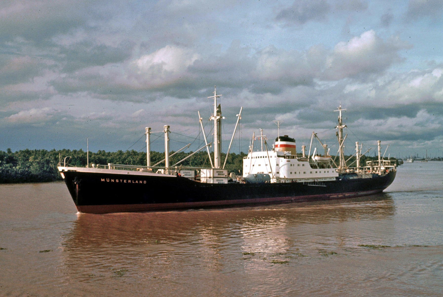 A color photo of a container ship on a muddy river in Thailand.  A tree-lined bank and cloudy sky are in the background. The ship's black hull is labeled Munsterland in white block capitals.  Several white containers are visible on the deck.  A smokestack with blue, white, and red stripes painted along the top sits on top of a white cabin area.