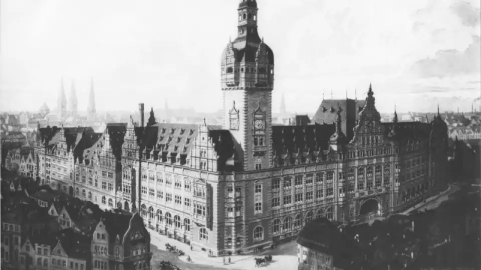 A black and white photo of a large neorenaissance-style building which takes up an entire city block, with smaller buildings in the same style in front of it across the street intersection.  A tower ascends from the corner at the intersection with a bell tower at the top.  Multiple pointed gables are placed along the roof above the entrances, which are spaced along  both visible sides of the building.