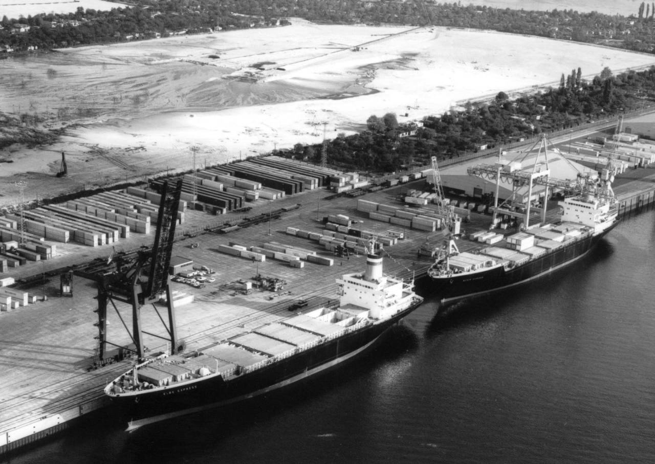 A black and white photo of a shipping dock.  Two container ships, the Weser-Express and the Elbe-Express, are pulled up next to it one in front of the other. A large flat area the size of several city blocks has been cut into the trees surrounding the dock, seemingly with nothing on it but dirt. In front of that, lines of shipping containers sit on two asphalt lots, separated by a road which passes through parallel to the river.  Two large cranes stand next to the ships.