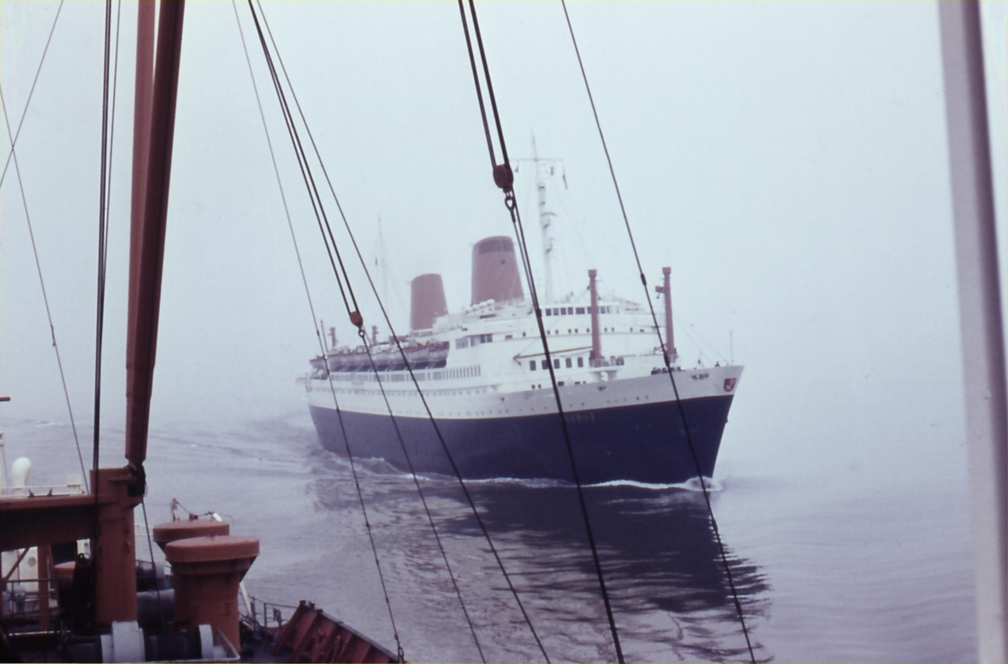 A color photo of the MV Bremen sailing out of the fog, moving toward the right of the frame as though to pass next to the viewer. The photo is taken from the deck of another ship, whose cables and orange-painted deck fittings ascend upward in front of the viewer.