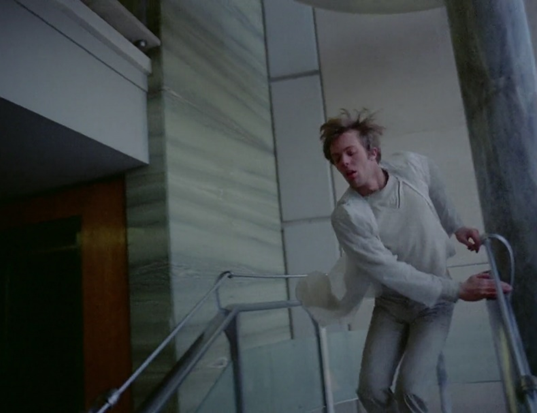 Max running down a flight of stairs dressed in all white and in a long white coat.