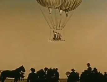 Long shot of cancan dancers on a balloon suspended platform with onlookers on the ground in shadow.