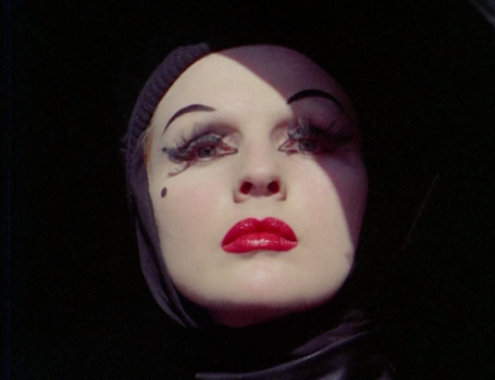 Close up on the face of an androgynous figure, in heavy pale makeup, and wearing a black hood
