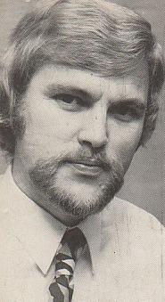A black and white author's headshot of a blonde white man with a close-cropped van-dyke beard, wearing a white dress shirt and striped necktie, looking at the camera with a neutral expression.