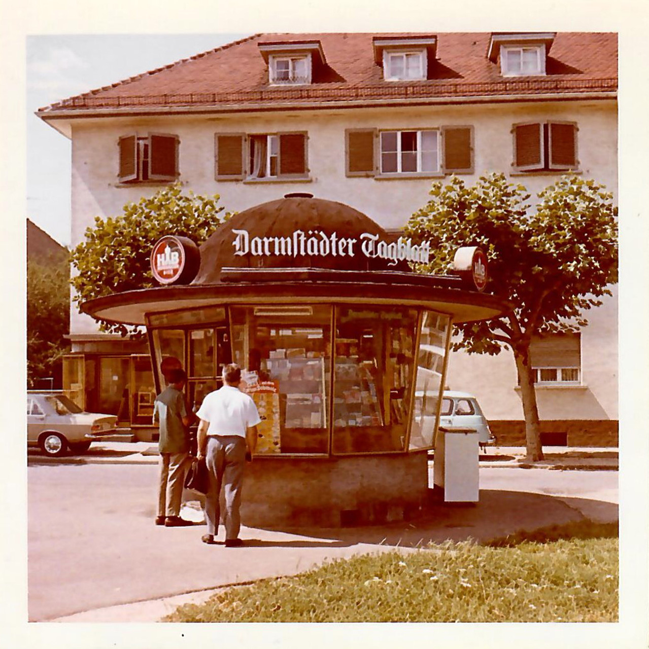 A faded color photo of a small round newsstand with windows all around the circumference and a bell-shaped roof that extends outward over the windows .  Two men wait in line at the open window. In the background, a three-story whitewashed building with a red roof and shuttered windows stands across the street.