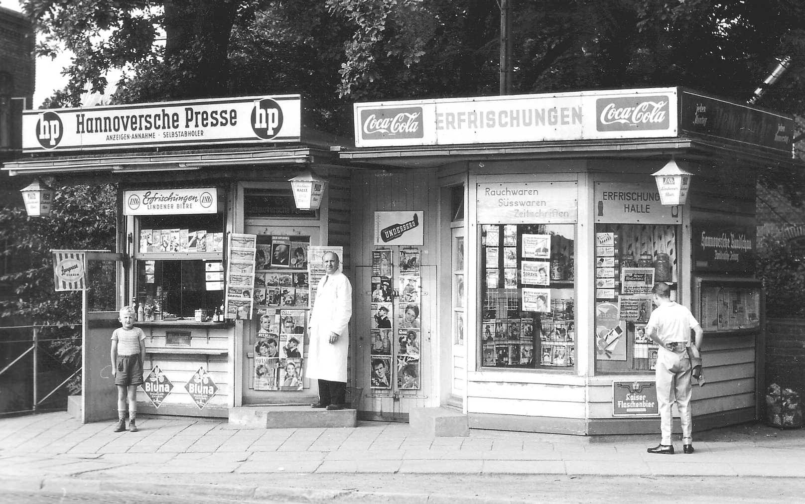 A black and white photo of a wooden-sided newsstand covered in advertising posters.  A man in a long white coat and a small boy in short pants pose on opposite sides of the newsstand window and door.  On the opposite side of the building, another man stands reading one of the advertising posters.