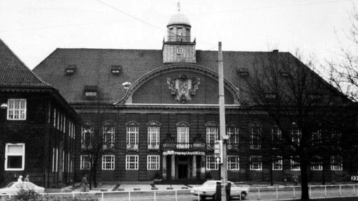 A black and white photo of a three-story Baroque revival-style building  taken from across the street.  Cars are passing in front of it. An arch at the center of the roof frames a large stylized heraldic shield.  A cupola above the arch has a small weather vane on top.